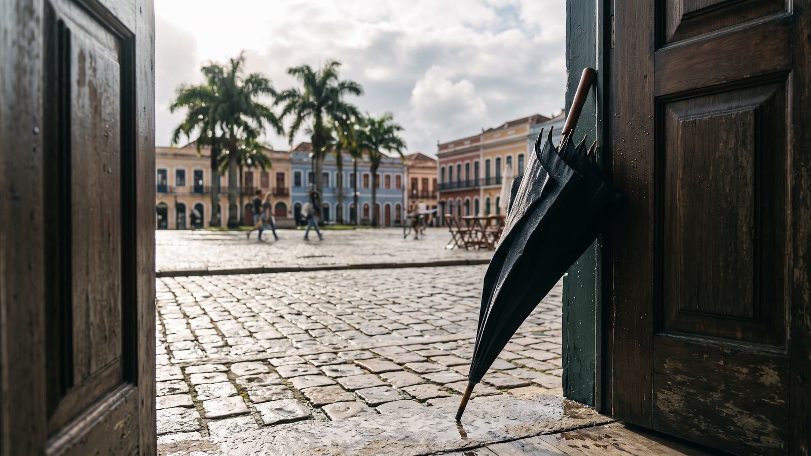 Paraguas mojado apoyado en la puerta de un edificio histórico mientras el sol vuelve sobre la plaza.
