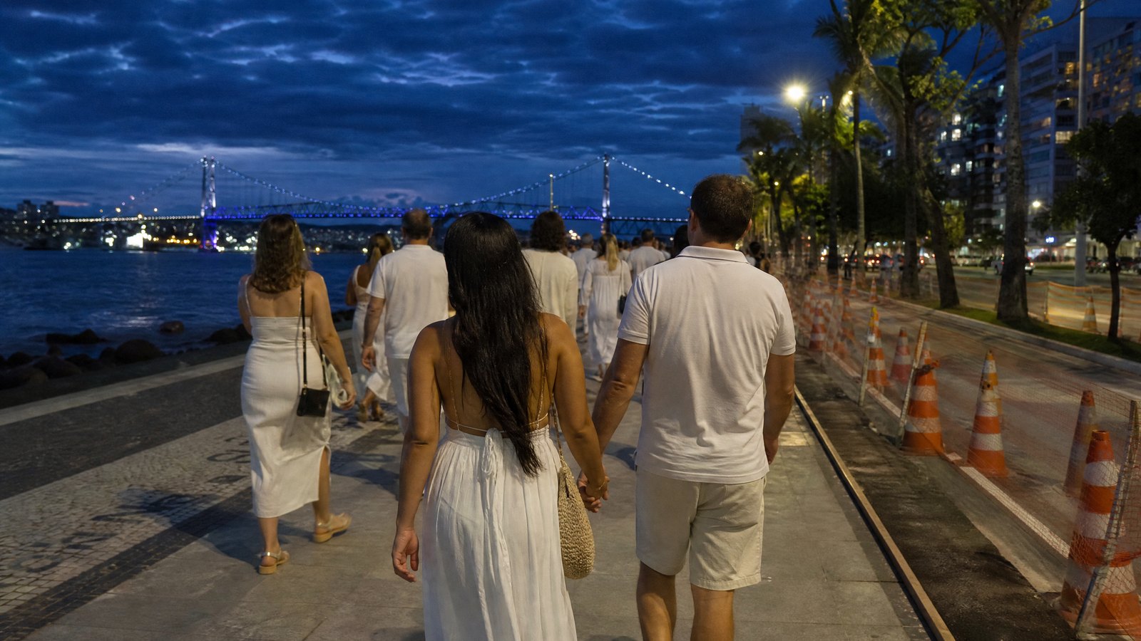 Gente vestida de blanco caminando por la Beira-Mar Norte al anochecer, con conos de obra y el puente Hercílio Luz iluminado al fondo